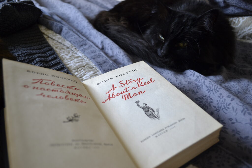 A black cat sleeps beside Polevoy's A Story of a Real Man. The book is open to the title page, which has red text and an illustration of an army pilot.