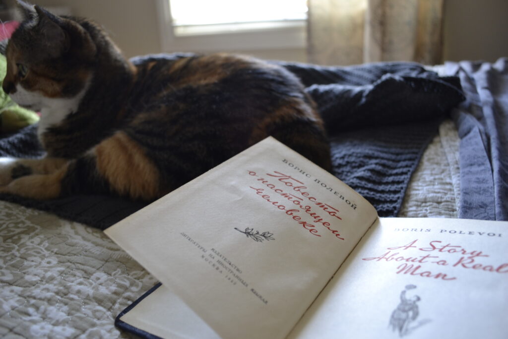 A calico tabby sitting beside the title page of Polevoy's A Story of a Real Man. The title in the book is red and there is an iullstration beneath it and the Russian translation on the left page.