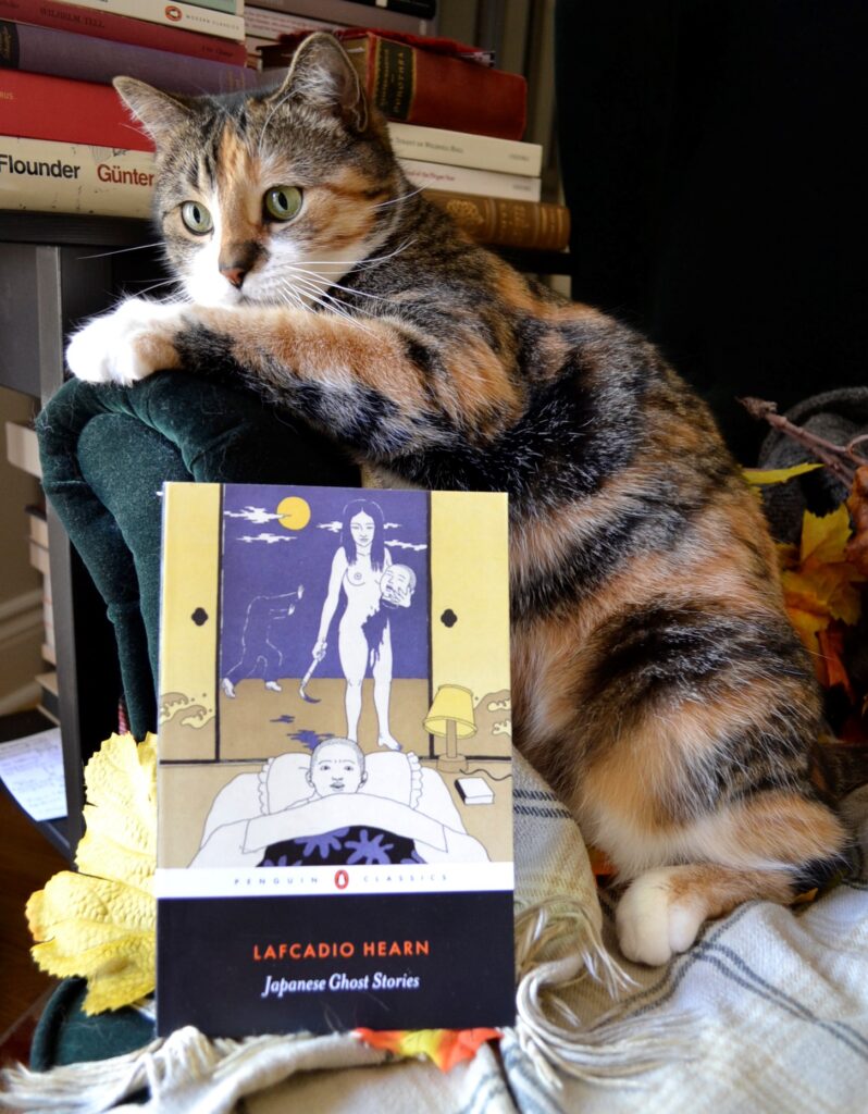 A calico cat sits in autumn leaves beside Japanese Ghost Stories.