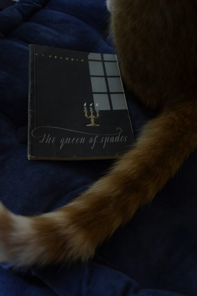 An orange tabby's tail wraps around the cover of The Queen of Spades.