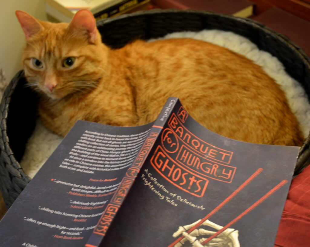 An orange tabby next to the cover of A Banquet of Hungry Ghosts