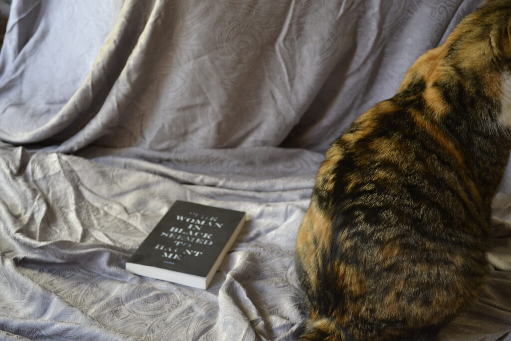 A calico tabby sits beside The Woman In Black.