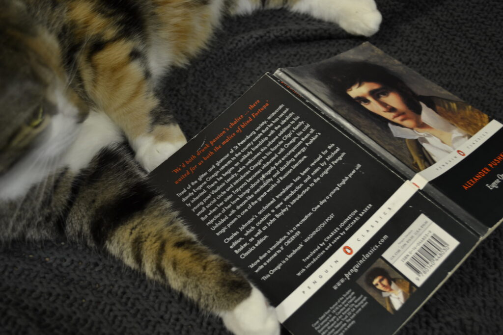 A calico tabby cat lounges beside a copy of Eugene Onegin.