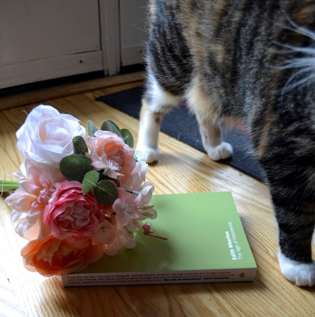 A calico tabby stands beside pink flowers and The Age of Innocence.