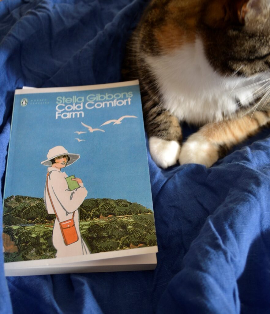 A calico tabby sits bside Cold Comfort Farm.