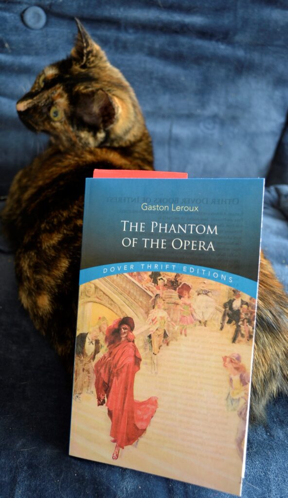 A tortoiseshell kitten sits beside The Phantom of the Opera.