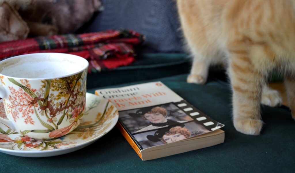 A teacup and orange paws beside Travels with My Aunt.