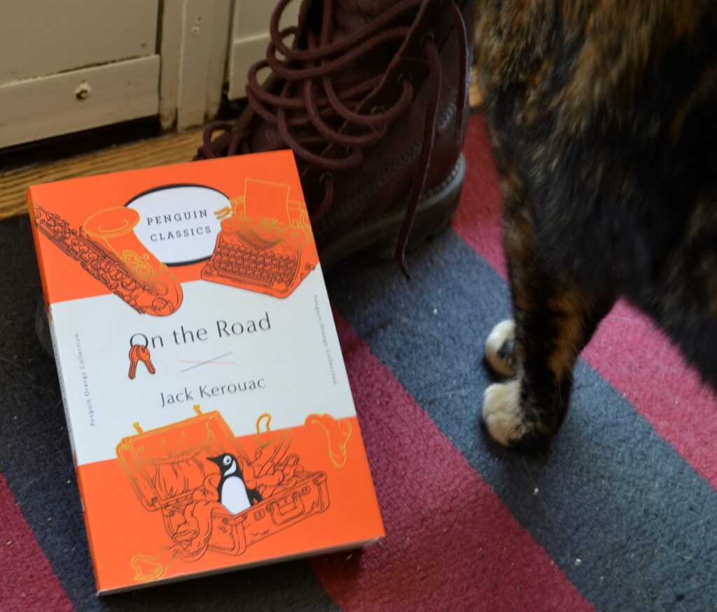 A tortoiseshell cat standing beside an orange copy of On the Road and a brown boot.