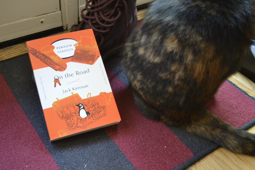 A tortoiseshell cat beside an orange copy of On the Road and a brown boot.