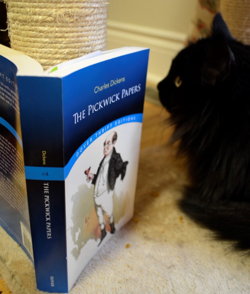 A black cat sits beside a copy of The PIckwick Papers.