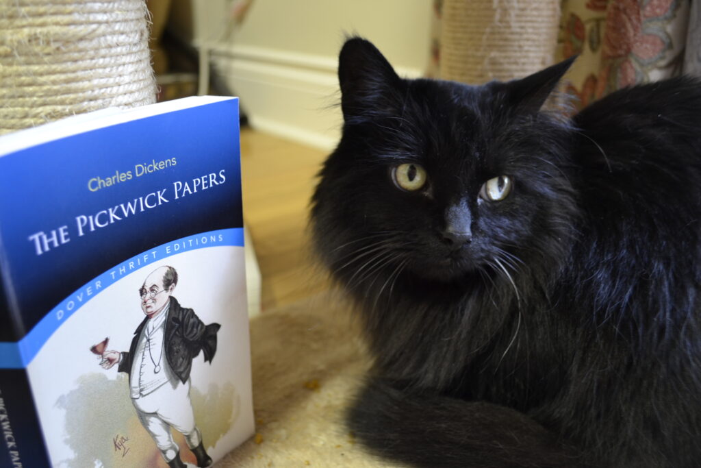 A black cat sits beside a copy of The PIckwick Papers.