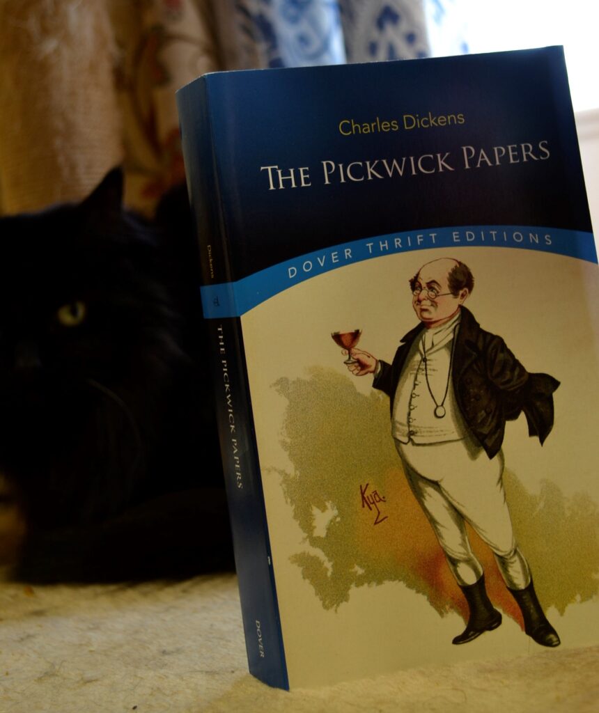 A black cat sits beside a copy of The PIckwick Papers.