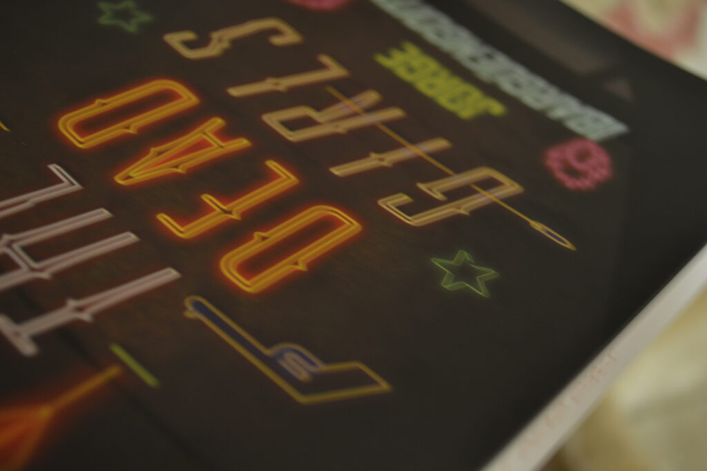 A close-up of a black cover with The Dead Girls written in neon lights.