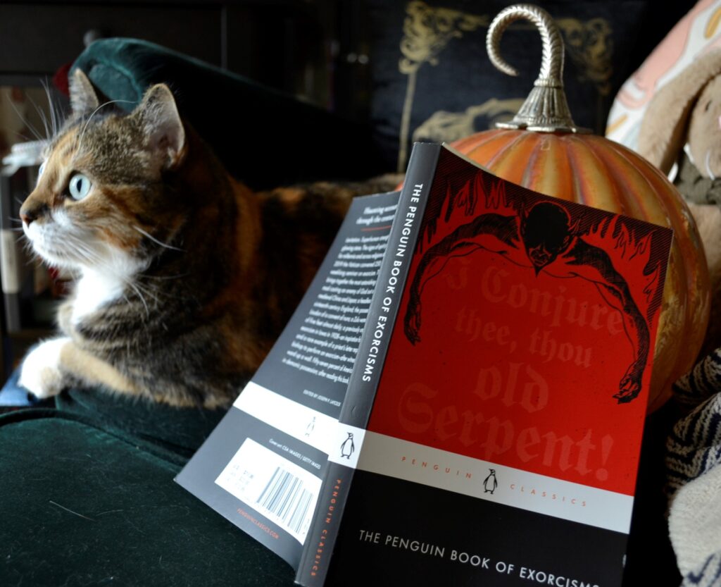 The Penguin Book of Exorcisms is spread over a glass pumpkin beside a calico tabby.