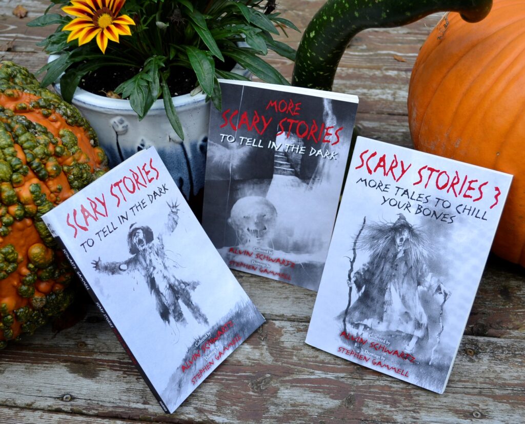 Three books with sketchy and eerie black-and-white illustrations are posed beside some pumpkins