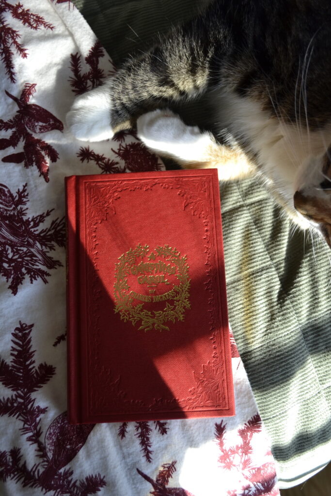 A calico tabby's paws rest beside an ornate red version of A Christmas Carol.