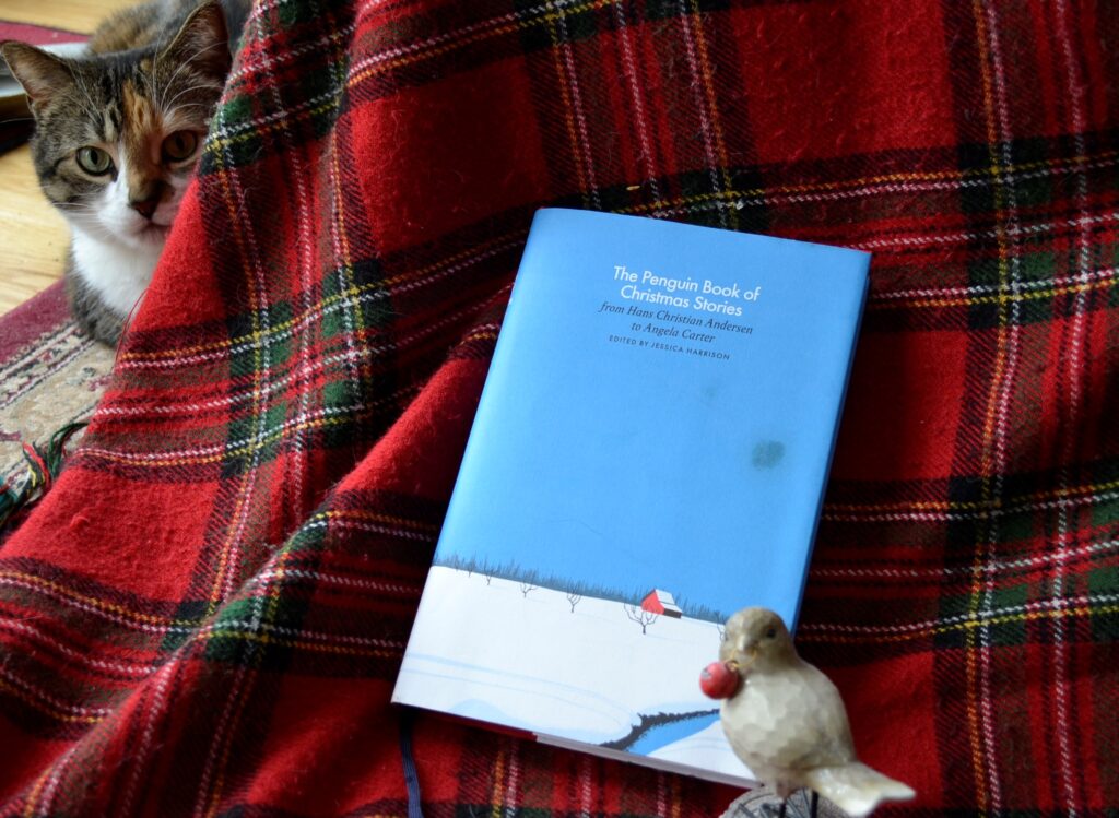 The Penguin Book of Christmas Stories lies on a red tartan with a cat behind it.