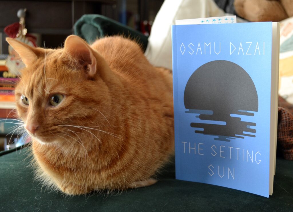 An orange cat sits beside a blue book called The Setting Sun.