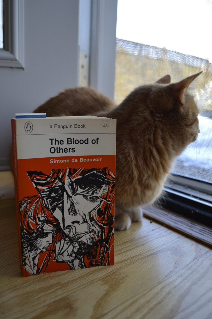 An orange tabby sits beside The Blood of Others.