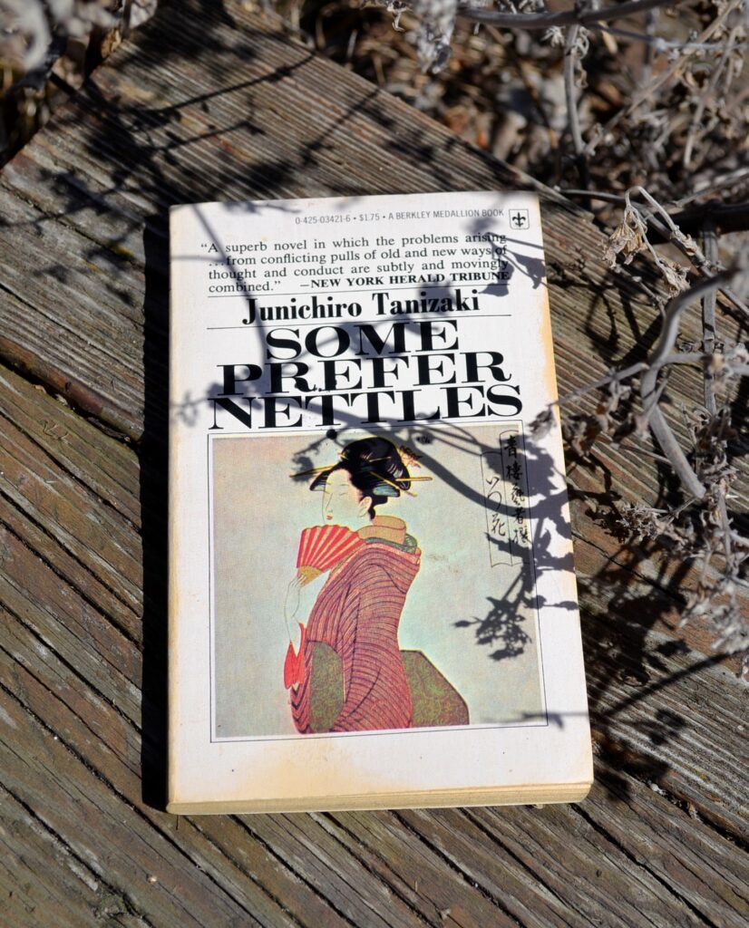 Beneath the spiny shadows of a dead plant, lays a worn paperback. The book features a woman in a kimono looking over a fan. The title is Some Prefer Nettles by Junichiro Tanizaki.