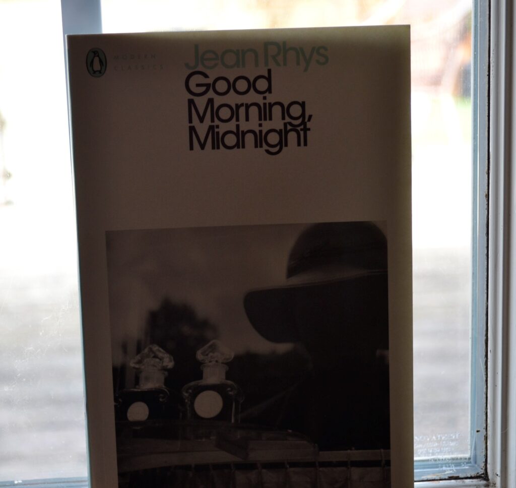 A white book with a black-and-white cover photo reads "Jean Rhys Good Morning, Midnight".