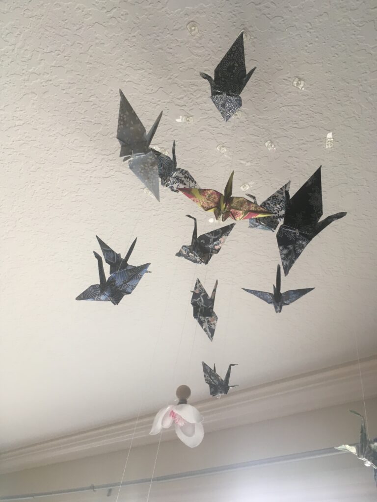 A handful of paper cranes in blue and white patterns and a white flower cloat in the air, suspended by thin string.