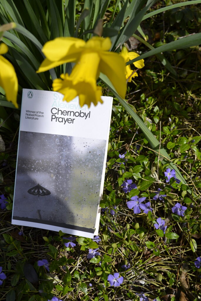 A daffodil obscures part of Chernoybl Prayer. The book is resting in perriwinkle.