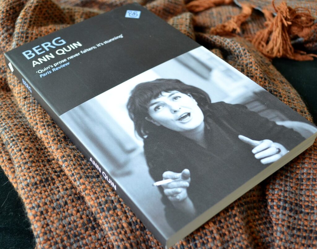 Berg by Ann Quin is a black book, featuring a black-and-white photograph of Quin on the front. Quin is smoking a cigarette and talking while gesturing with her hands.