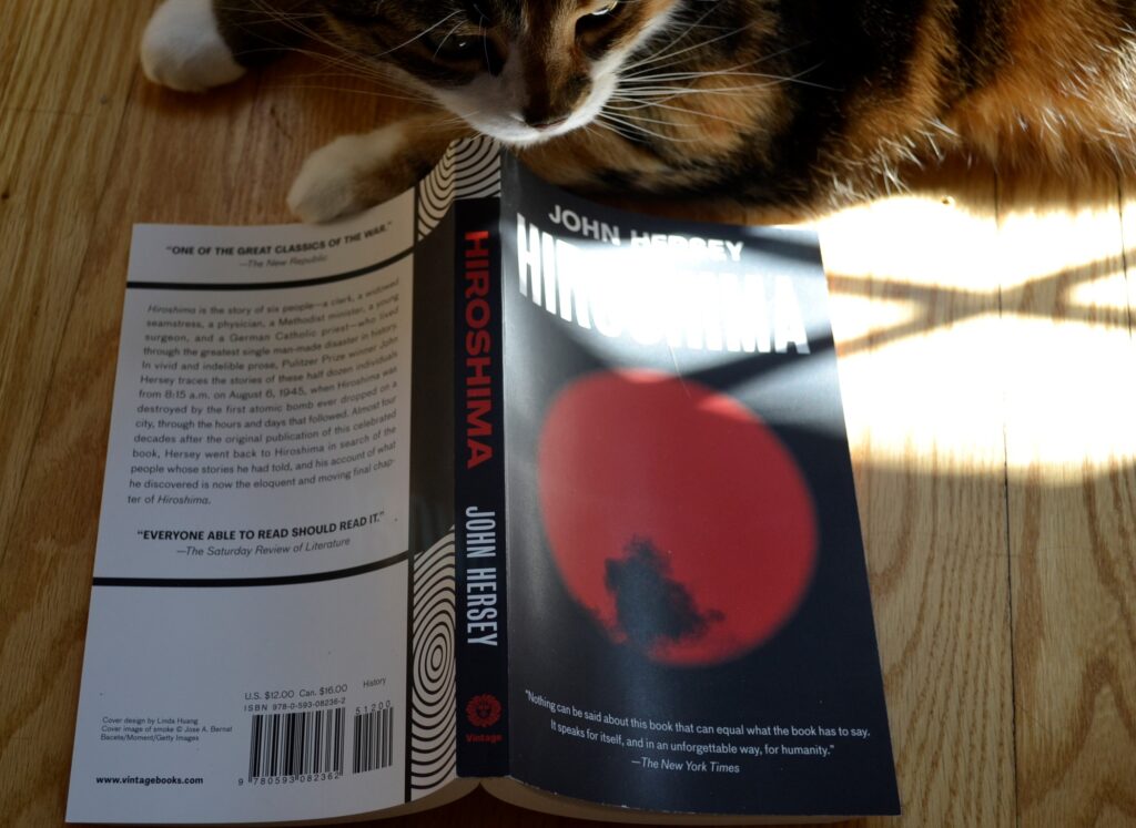 A calico tabby sniffs a spread out book. The book is Hiroshima.