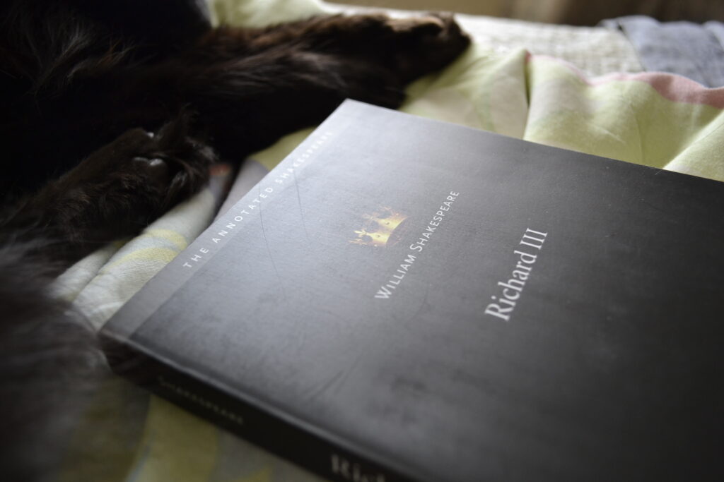 Black paws curl beside a black book that features only a small crown and the words 'William Shakespeare' and 'Richard III'.
