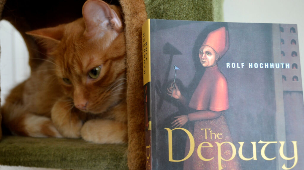 An orange tabby cat frowns beside the edge of a book. The book clearly reads 'Rolf Hochhuth, The Deputy'.