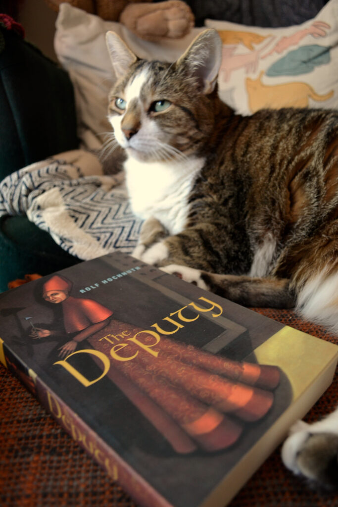 A tabby cat looks off into the distance, her paws curled around a copy of Rolf Hochhuth's The Deputy.