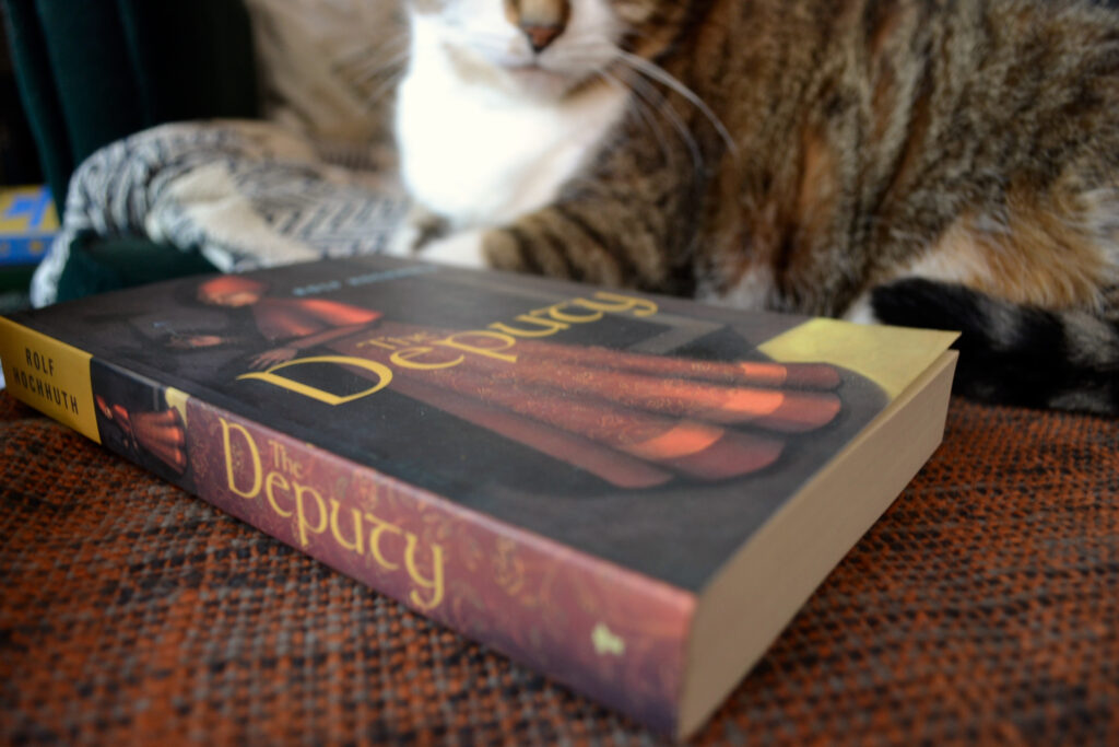 The spine of a book reads The Deputy. Behind it, a tabby cat lounges.