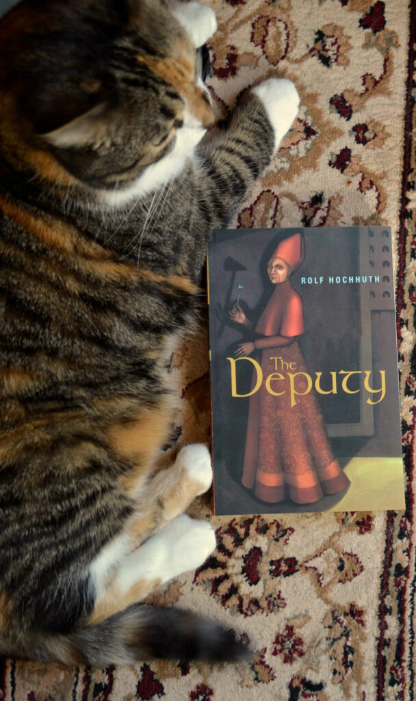 A calico tabby lies beside a book titled The Deputy. The cover features a painting of a pope.