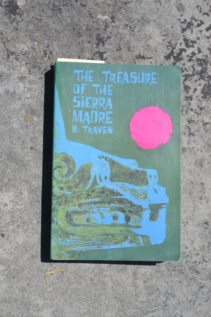 A blue and green book with a picture of an aztec stature and a red sun is titled The Treasure of the Sierra Madre.
