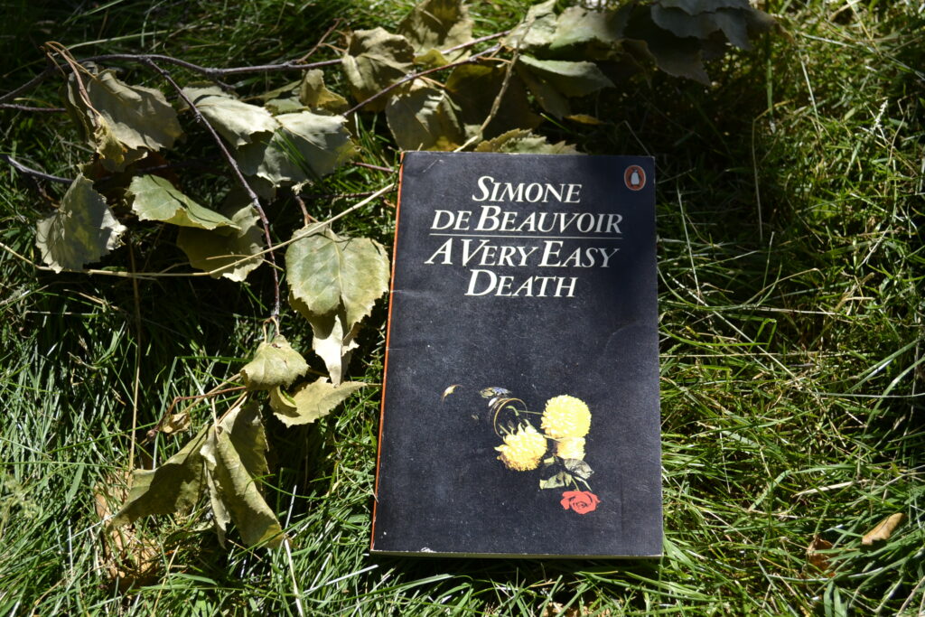 A book with a black cover lies in the grass. The white font reads 'Simone de Beauvoir | A Very Easy Death' and the one small image is of a falled vase of flowers.