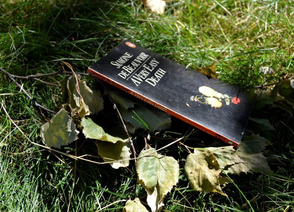 A Very Easy Death lies in the grass. The cover is black with only a small image of a tipped-over vase of flowers. The spine is orange.