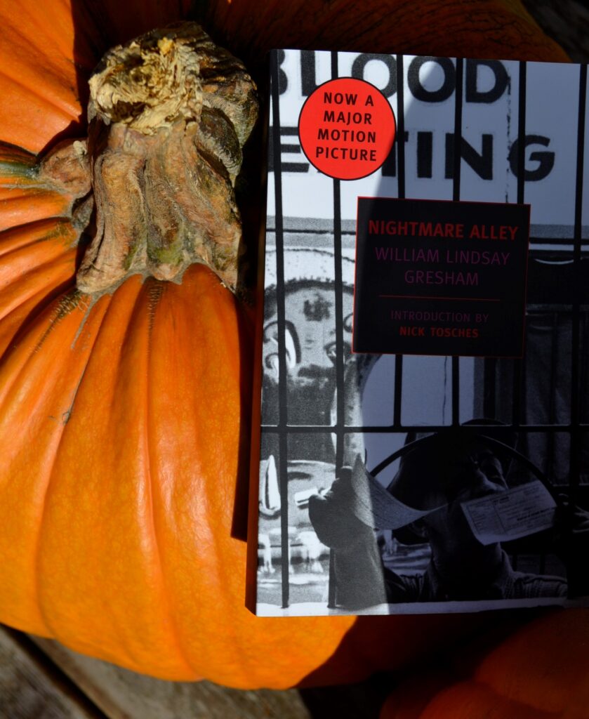 A copy of Nightmare Alley by William Lindsay Gresham sits on a pumpkin. A black-and-white cover shows a carnival ticket booth.