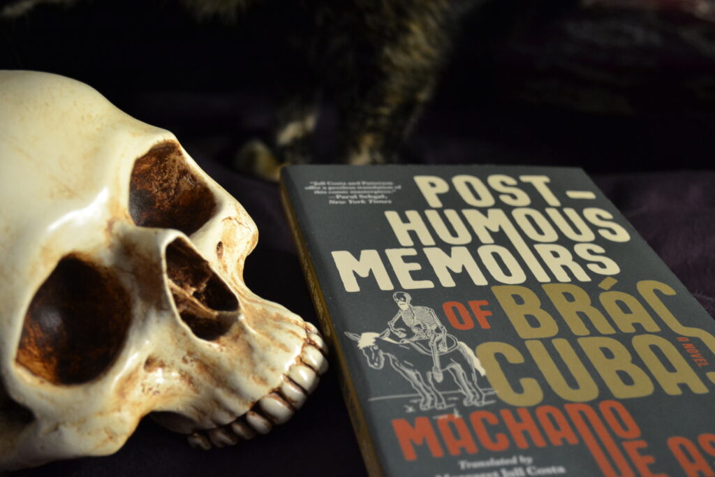 A skull sits beside a copy of The Post-Humous Memoirs of Bras Cubas. On the cover is a drawing of a skeleton riding a donkey.