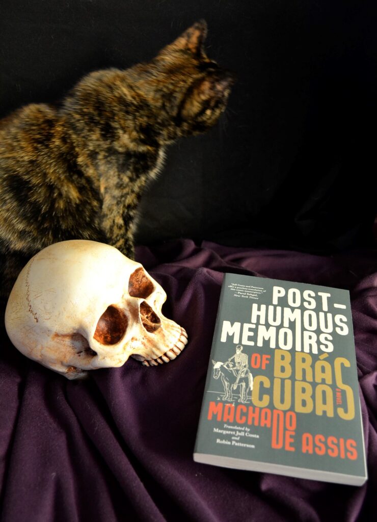 Machado de Assis' Post-Humous Memoirs of Bras Cubas sit on a purple cloth beside a tortie cat and a skull.