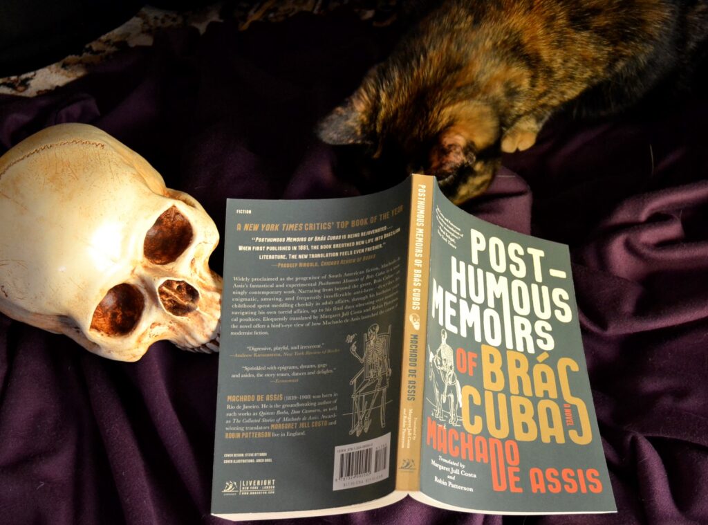 A skull and a tortoiseshell cat sit beside a copy of The Post-Humous Memoirs of Bras Cubas.