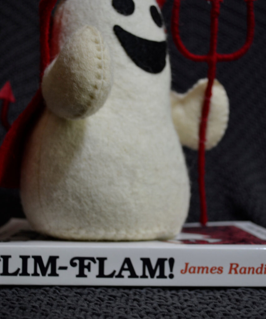 A stuffed ghost figure is dressed as a devil and smiling widely. It sits on a copy of James Randi's Flim-Flam!