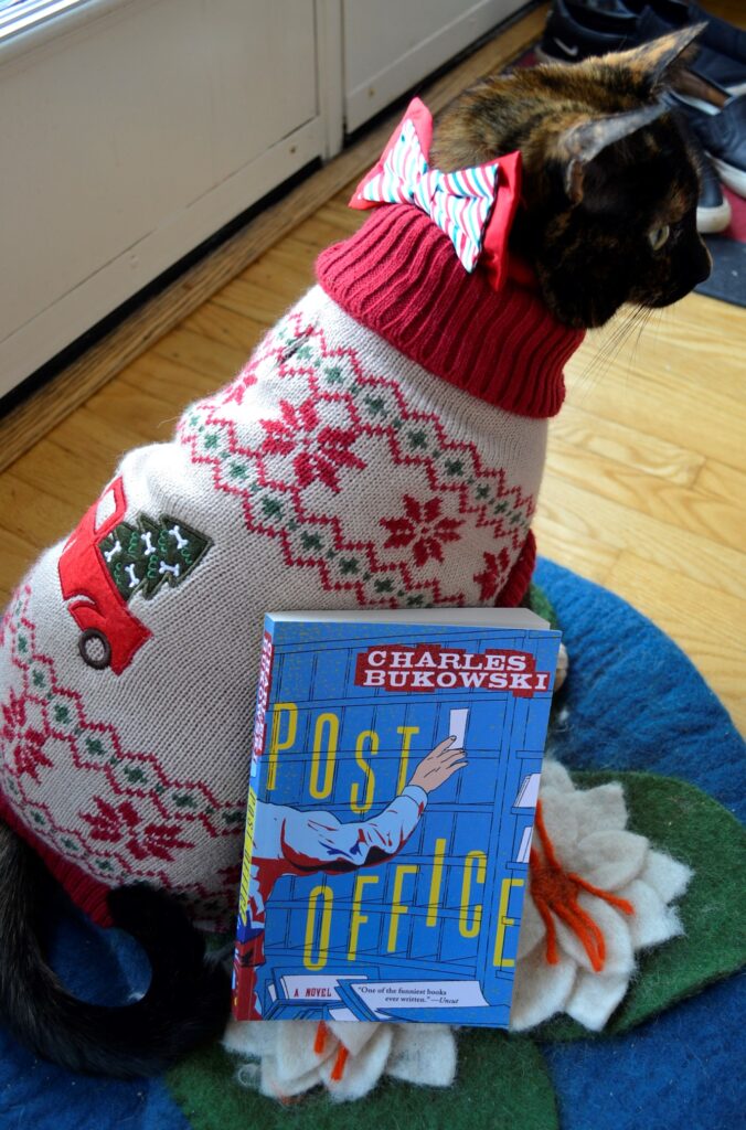 A copy of Post Office leans against a cat that is wearing a sweater.
