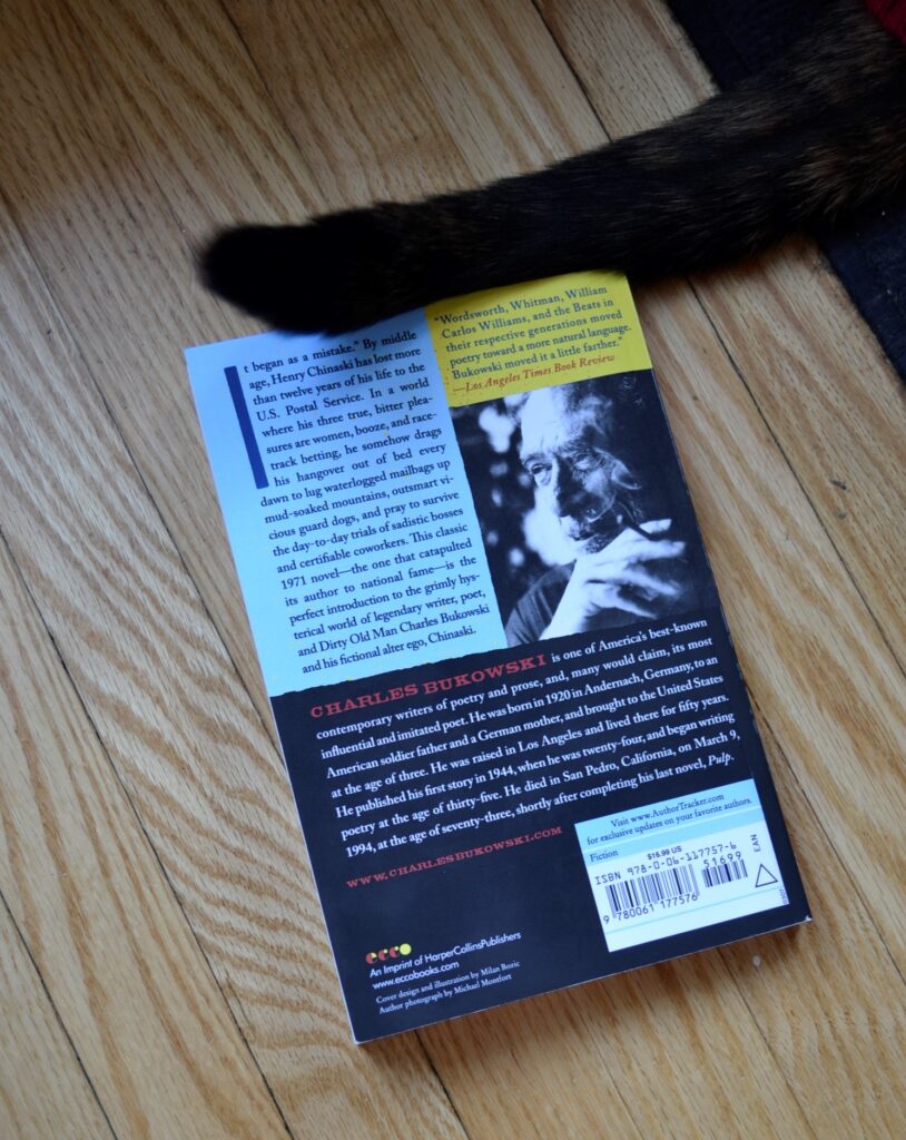 A cat's tail brushes the top of a book. The book's back cover is up, with a picture of Charles Bukowski in black-and-white on it.