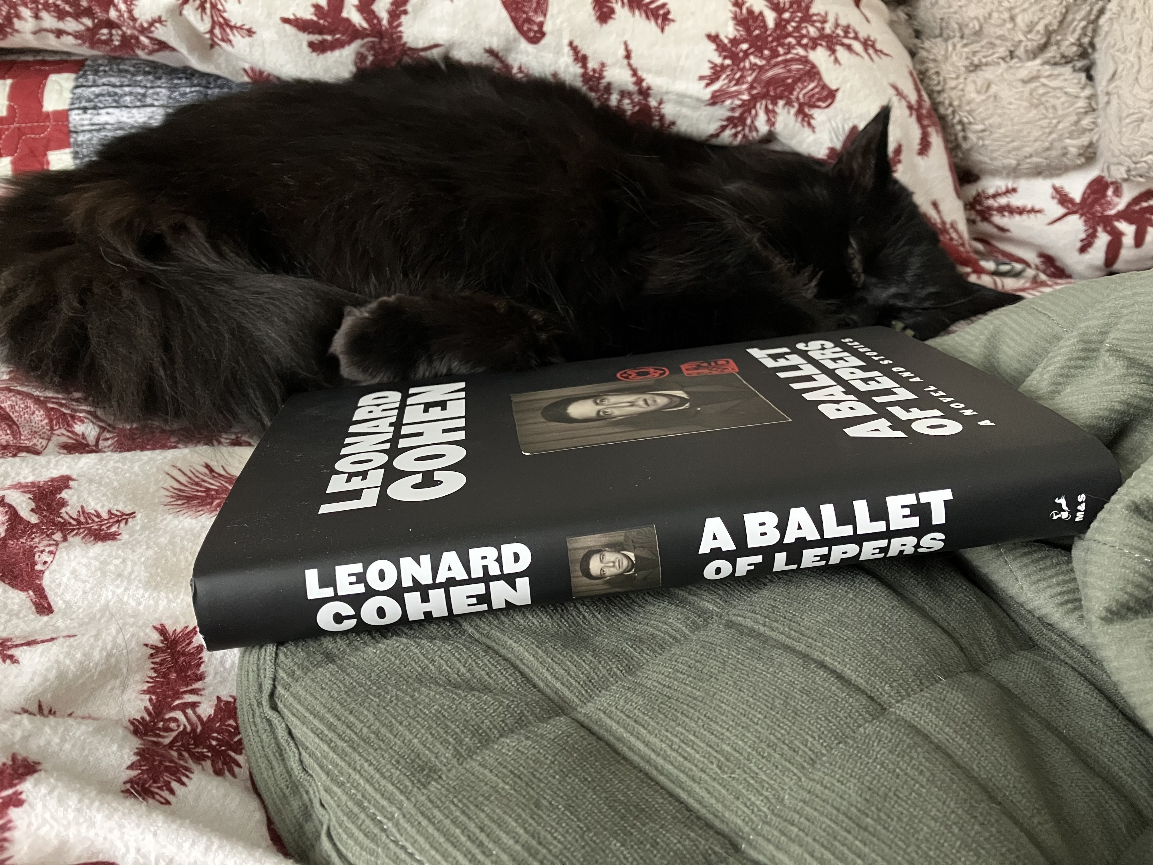 A black cat sleeps behind Leonard Cohen's A Ballet of Lepers. The book is black with white block font and a passport photo of Cohen on the cover.
