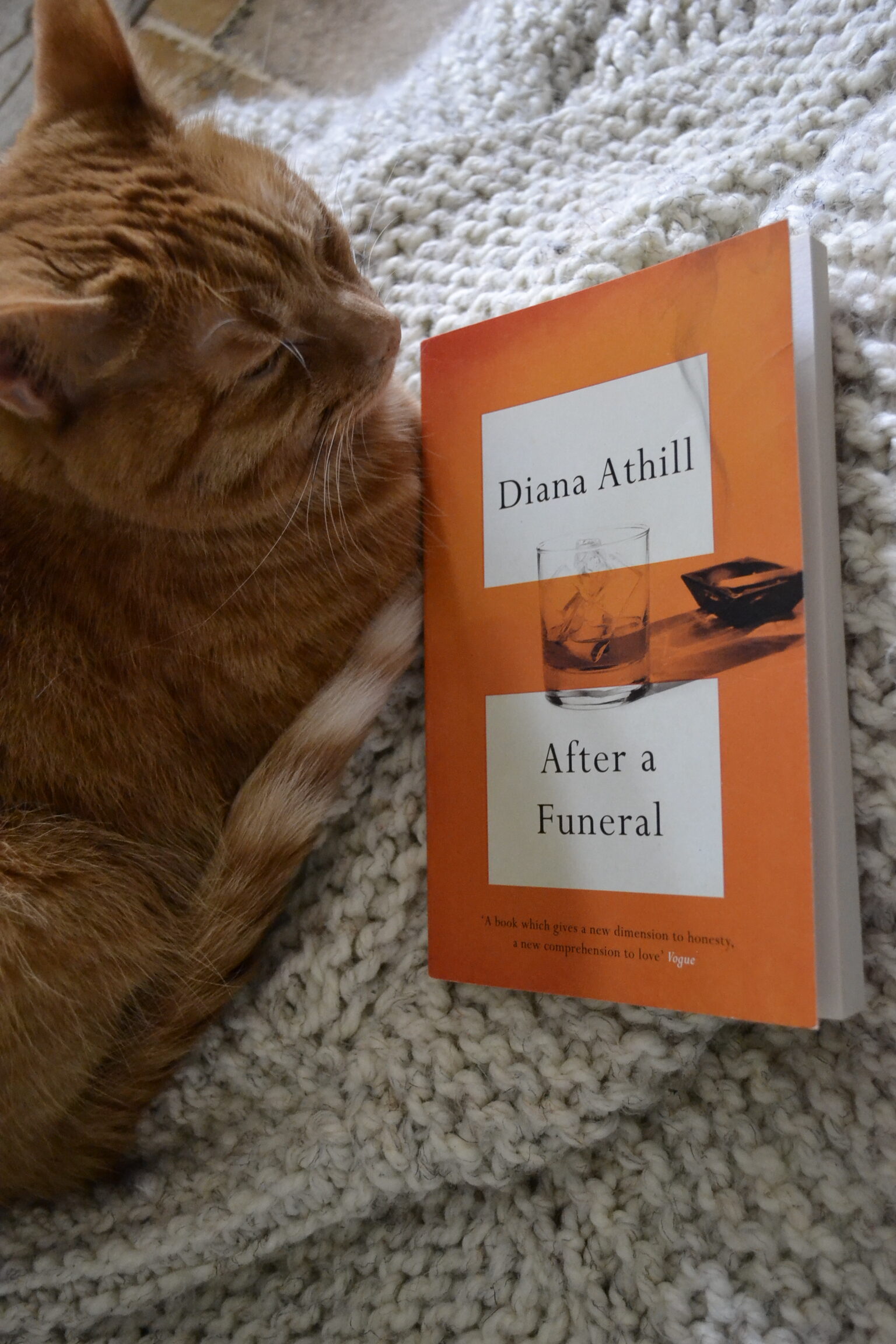 On a white blanket, an orange cat sleeps beside an orange book. The book is After a Funeral by Diana Athill.