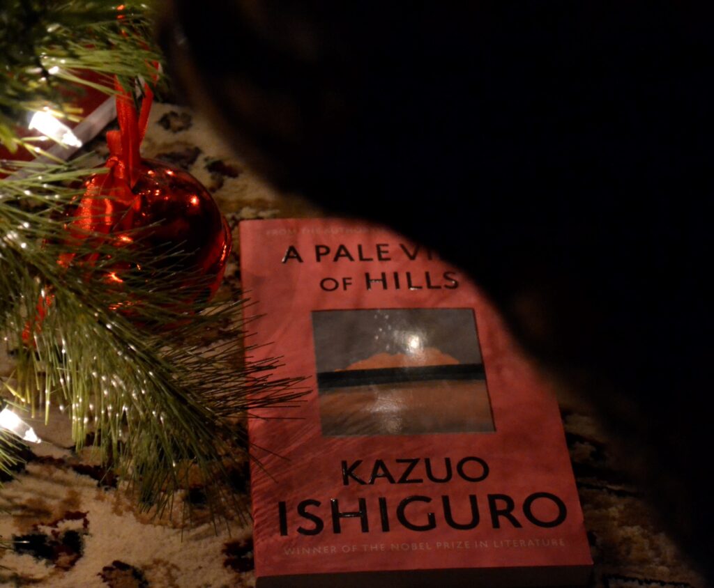 A cat stands over a pink book. The book is A Pale View of Hills by Kazuo Ishiguro.