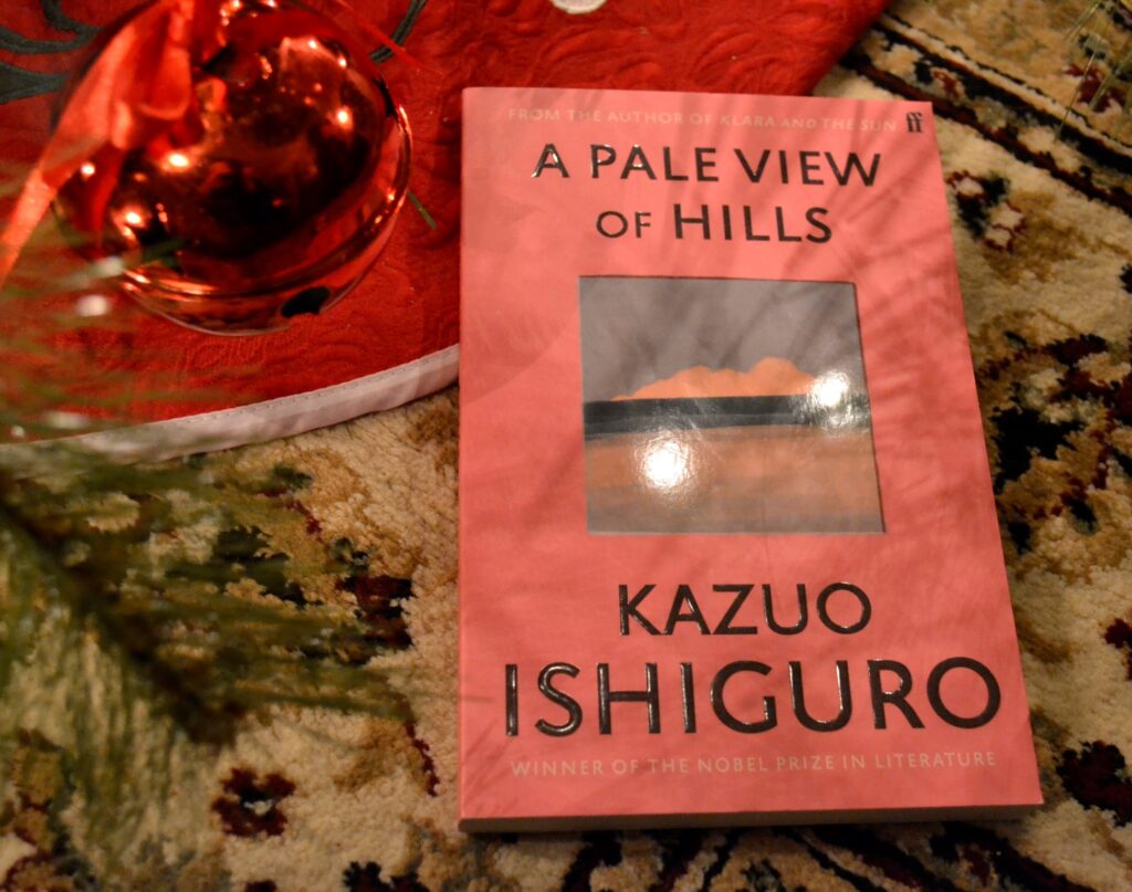 A red bell hangs beside a book. The book has a pink cover with an image of a sunset in the middle. It is A Pale View of Hills by Kazuo Ishiguro.