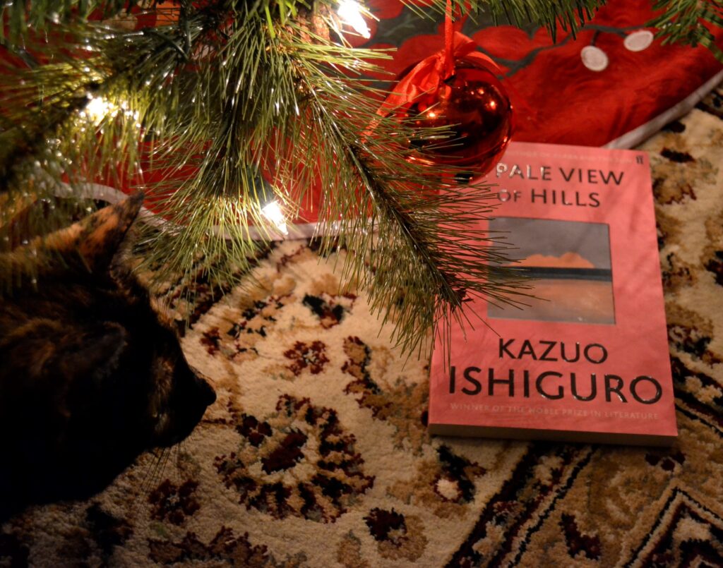 Beneath a Christmas tree, a cat looks at a pink book with a sunset on the cover. A red bell ornament hangs above them.