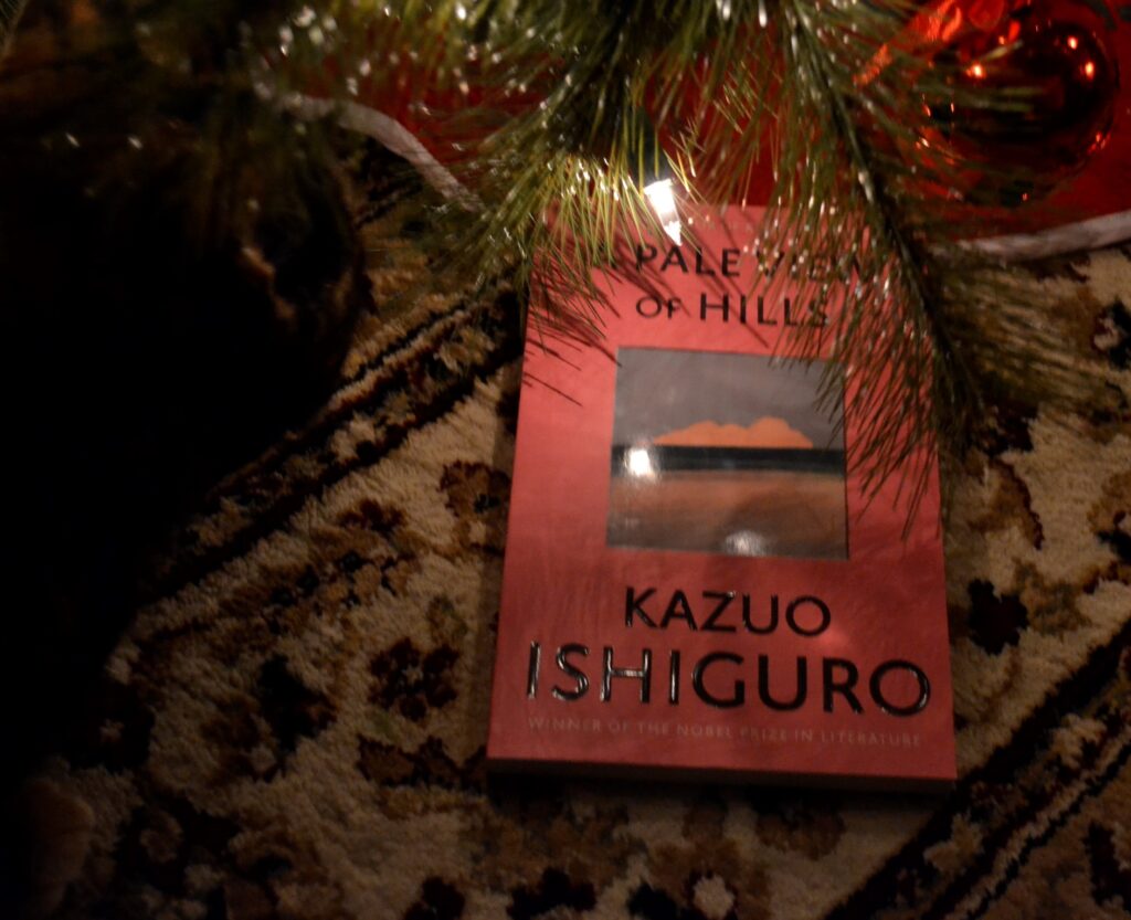 A cat sits beside a book beneath a Christmas tree. The book is Kazuo Ishiguro's A Pale View of Hills.